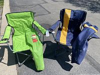 Two camping chairs set outdoors on pavement, one green and one navy blue with yellow stripe, each with matching carry bags.