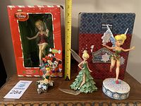 Four Disney Tinkerbell figurines displayed on a wooden surface with a measuring tape showing about 8-10 inches height, and original boxes behind.
