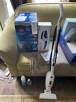 Photo showing Bissell SpotClean ProHeat box, BrushSteam steamer box, white Featherweight vacuum on couch and floor with cleaning liquids