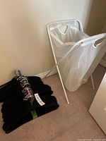 Photo showing the white laundry basket with metal frame and fabric bag next to a stack of black velvet hangers on a carpeted floor.