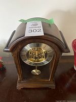 Wooden mantle clock with curved arched top and visible internal brass clockwork and pendulum through front glass panel. Clock showing time with Roman numerals on dial.