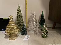 Wide view of six different Christmas tree decorations including two clear glass trees, two green glittered trees, one green ceramic with red berries, and one gold glittered spinning tree topper.