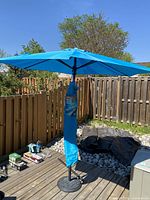 Ocean Blue rectangular deck umbrella fully open, mounted on a weighted metal base on a patio deck with surrounding fence and garden in background.