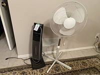 Wide view showing the black tower heater and white standing fan positioned side by side against a beige wall on tile floor with patterned rug.