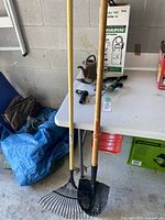 Photo showing metal leaf rake, metal garden spade, and wooden handles standing against a concrete wall.