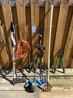 Wide view of garden tools including edger/trimmer, extension cord, cultivator, loppers, hand rake leaning against wooden fence on deck