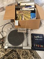 Kenwood turntable with dust cover, box of assorted vinyl records, Genesis book laid out on carpet