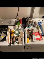 Painting supplies including brushes, paint rollers, paint trays, masking tape, paint cans, gloves, and other supplies on top of washing machines.