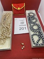 Overview photo showing two pearl necklaces in Jackie Orr boxes (one cream, one grey), two pairs of pearl stud earrings, and a 14k gold pearl pendant on a red background.