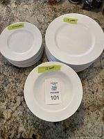 Stack of white porcelain dinner plates (13 pieces), lunch plates (12 pieces), and bowls (12 pieces) arranged on a countertop with handwritten labels indicating quantities.