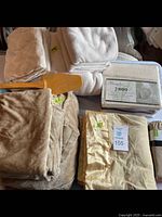 Overview of all bedding items including plush blankets, bamboo sheet set in box, satin bordered cream sheet, and wooden bed lifters visible