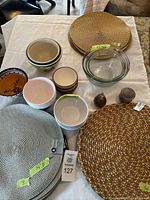 Display of various bowls including glass Anchor Hocking mixing bowls, ceramic bowls in assorted colors, white plastic bowls, glass pear-shaped décor items, and a covered stone container along with round woven placemats on a white tablecloth.