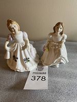Two Royal Doulton figurines named February and Lynsey displayed side by side on a grey surface against beige wall, showing front view.