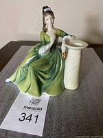 Front angled photo showing full figurine seated with dress flowing and hand resting on column holding bouquet.