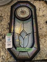 Full view of the stained glass wall clock showing iridescent glass panels and black frame with dimensions (10"w x 2"d x 22"h).