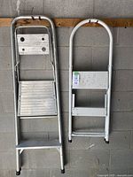 Two aluminum step ladders hanging on wall, one larger with a platform step, one smaller with two steps and white coating.