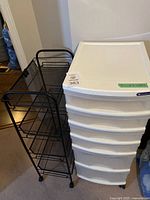White plastic storage cart with five drawers and black metal wire storage cart with shelves together in a room corner.
