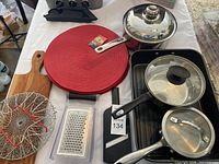 Photo showing circular red placemats, three metal pots with glass lids, two frying pans with lids, a rectangular roasting pan, a metal wire basket, and a flat metal grater with plastic cover.
