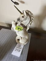 Unicorn figurine sitting with curled tail, showing white textured fur, gold horn, and blue crystal necklace.