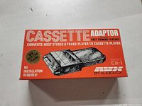 Red box packaging labeled 'CASSETTE ADAPTOR' with product image, highlighting 'No installation required' and 'New Auto Stop Feature'. Model CA-1 by Audiovox.