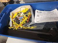 Blue container holding numerous black plastic slot car track pieces, yellow plastic guardrails, wired controllers, two slot cars, and an instruction sheet.