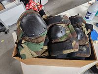 Box containing woodland camouflage colored military knee and elbow pads with black hard plastic protective caps, straps visible.