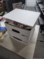 White archival Bankers Box filled with art supplies including paint markers and papers, photographed from above on a table.
