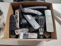 Box full of assorted remote controls, viewed from above, showing multiple units stacked inside worn cardboard box.