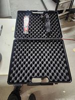 Open black hard shell carrycase with egg crate foam inside and a plastic sealed accessory pack inside the top foam