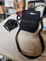 Photo showing the black Chinon Genesis III camera next to a black carrying case with shoulder strap on a white surface.