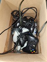 Top view of the box containing several wired game controllers including Xbox 360 and original Xbox controllers mixed with PS2 controllers, showing their condition and cables tangled inside the box.