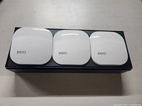 Three white square-shaped Eero mesh WiFi units with rounded edges on a black box.