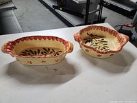 Two ceramic ovenproof dishes on white surface; both cream with painted olive branches and scalloped rims in reddish brown.
