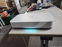 White HP Tango printer placed on a table with blue indicator glowing, showing front side with power cable connected and paper tray.