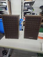 Two rectangular Marantz speakers with brown foam covers having a square grid pattern, placed on a white table.