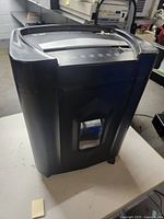 Black Pen+Gear paper shredder seen from front and side, placed on a white table. Rectangular bin with window visible.
