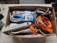Box containing various power strips and extension cords, some packaged new, others loose and coiled