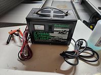 Front and top view of black metal Schauer tri-power charger with attached cables and clamps, showing branding, model info, and ammeter.