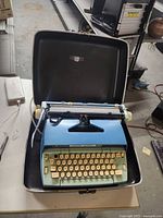 Blue Smith-Corona Coronet Electric 12 typewriter open inside black carrying case on floor, showing keyboard, platen roller, and typewriter internals.