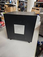 Front view of Dynaudio Solo Cinema Studio subwoofer showing black cubical design with front metal logo plate