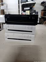 Front view of three stacked Nintendo Wii consoles, two white units on bottom and one black unit on top, showing disc drive slots and front power buttons.