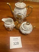 Top angle photo showing the entire tea set of three pieces: teapot, creamer, and sugar bowl with lids and gold ornamental designs, placed on a wooden table.