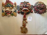 Three vintage Tibetan masks made of wood displayed on a white surface. The central mask has an ornate hanging wooden ornament.