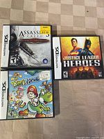 Front of three Nintendo DS game cases: Assassin's Creed, Justice League Heroes, Yoshi's Island.