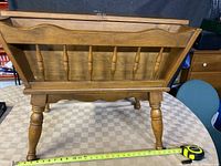 Front view of the solid wood antique magazine rack showing its curved top edge and turned legs with a measuring tape on table.
