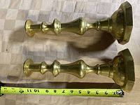 Pair of brass candlesticks lying on a surface with a measuring tape showing length approximately 11 inches.