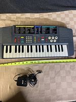 Full view of the Optimus MK420 keyboard showing keys, speakers, control panel, and power adapter below keyboard with cable coiled.