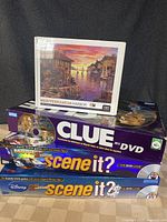 Puzzle with Mediterranean Harbor image on box, unopened in shrink wrap, shown stacked on top of board games including Clue DVD and Scene It?