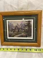 Framed Peter Dobson print artwork showing a cottage with warm lighting at dusk, with measuring tape showing frame width around 10-12 inches.