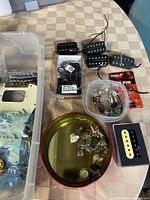 Photo showing pickups including black and cream humbuckers, a small box labeled Ibanez guitar humbucker, assorted tuning parts and knobs in a round container, plus plastic pickguards and metal bridge in plastic bin.